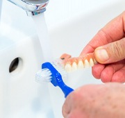 A person using a denture brush to clean their dentures in the sink
