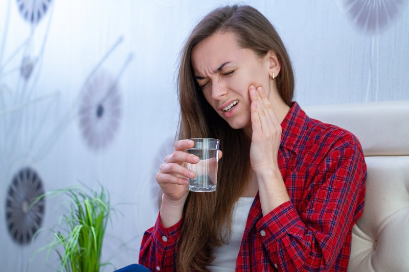 a young woman holding a glass of water in one hand and touching her cheek in pain with the other hand